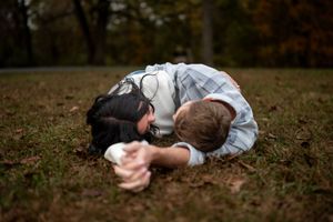 Two people in white shirts embrace while lying on autumn grass in a park setting.