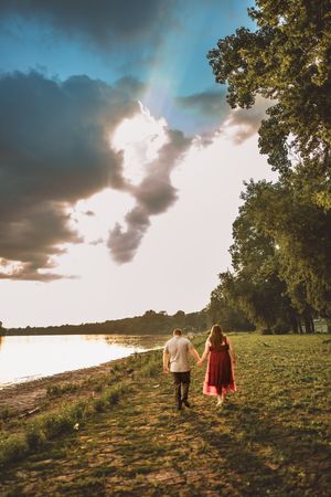 Two people walk hand in hand up a grassy hillside during golden hour with dramatic clouds overhead.