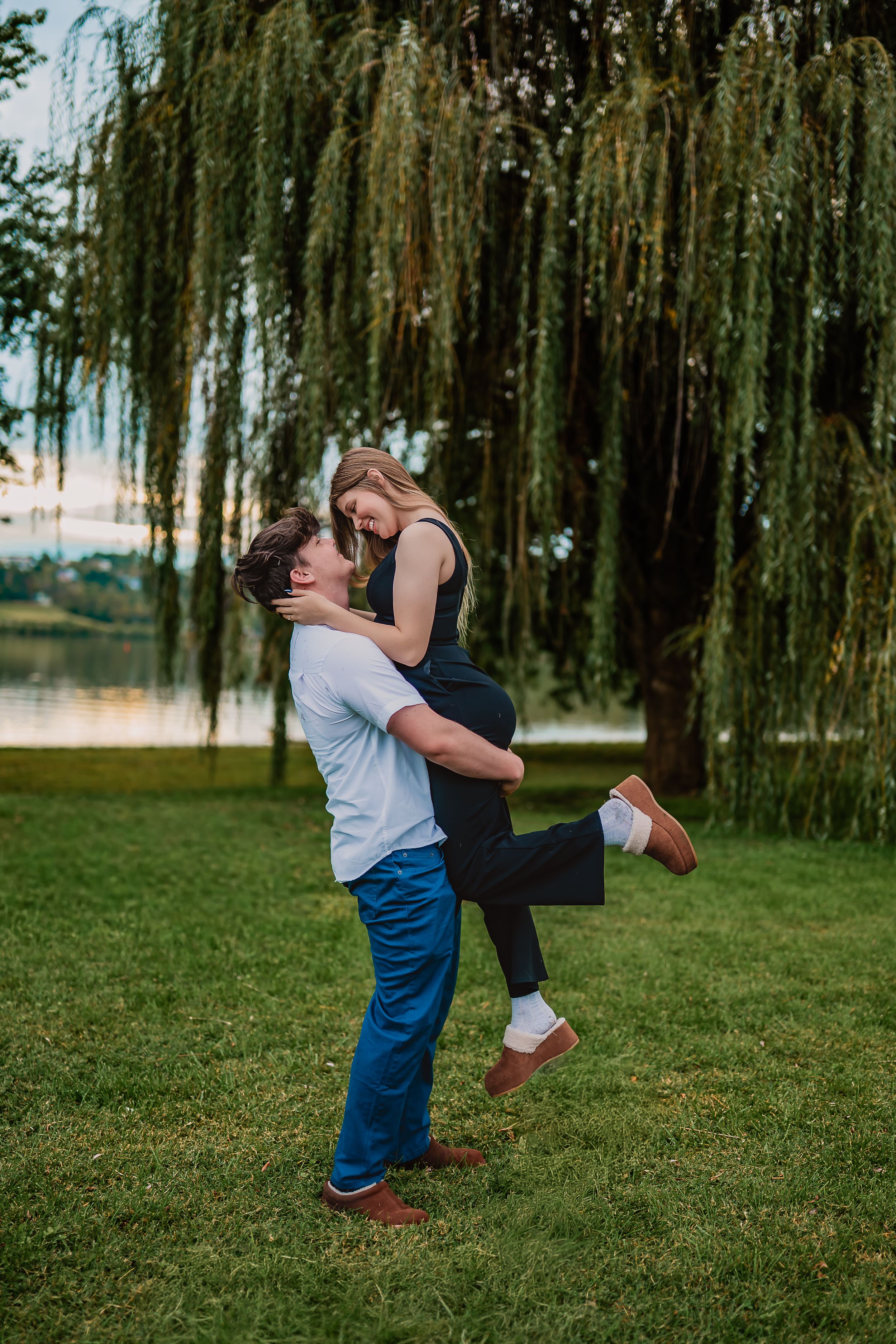 A couple dances playfully on a grassy lawn beneath drooping willow trees in a romantic outdoor setting.