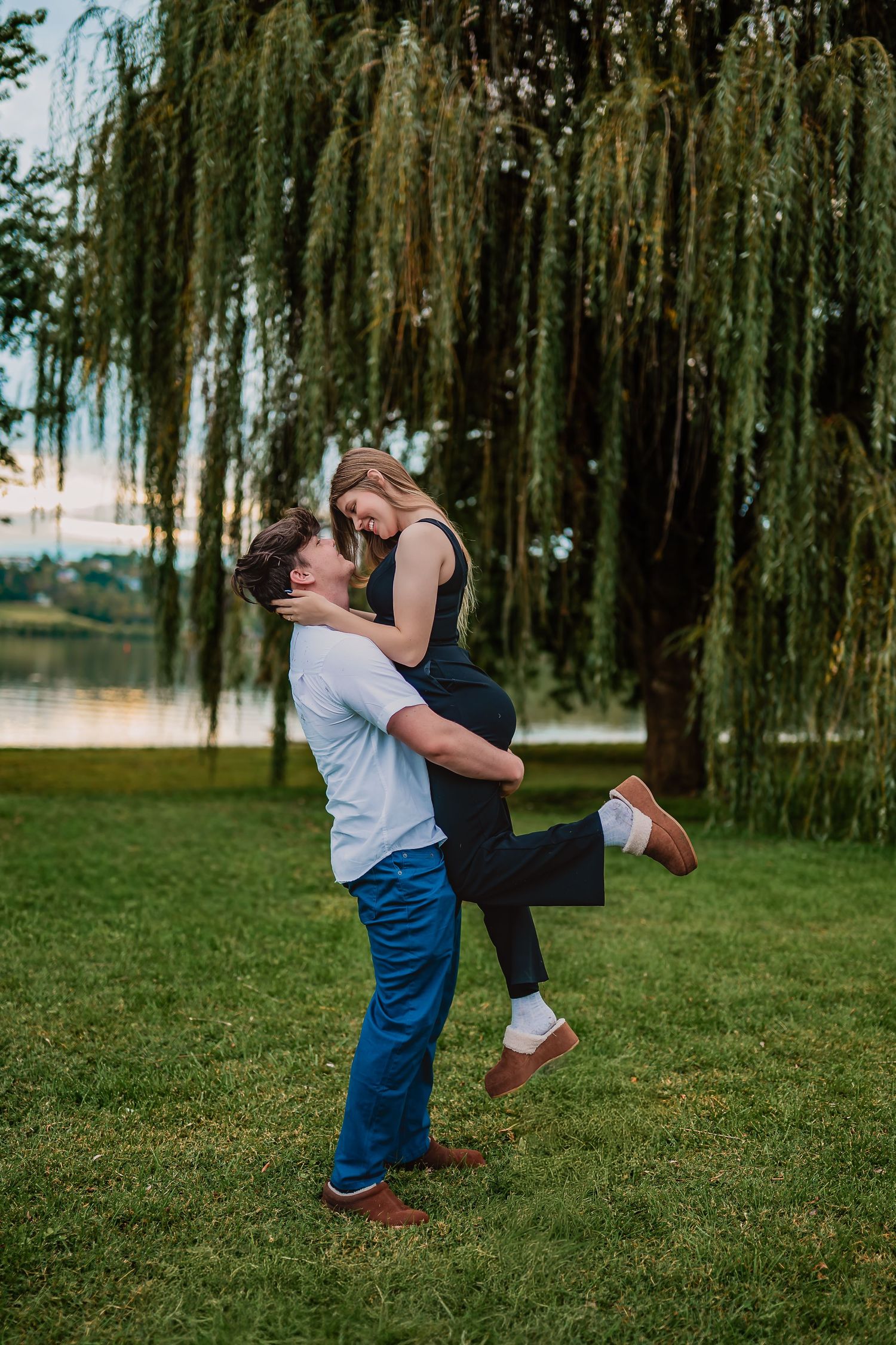A couple dances playfully on a grassy lawn beneath drooping willow trees in a romantic outdoor setting.