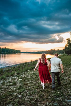 A couple walks along a rocky shoreline at sunset under dramatic blue storm clouds.