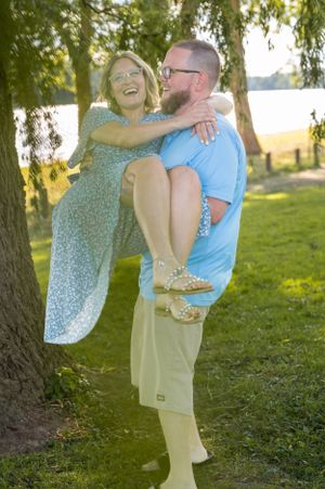 Playful couple having fun with piggyback ride under willow trees by lake.