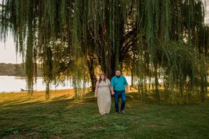 A couple walks together beneath weeping willow trees along a lakeside path at sunset.