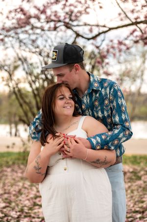 Couple embracing under cherry blossoms with man in baseball cap and blue patterned shirt.
