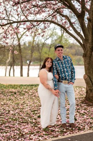 A couple poses under blooming cherry blossoms by a lake with pink petals scattered on the ground.
