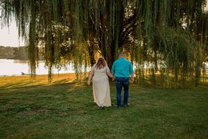 A couple walks beneath weeping willow trees along a sunlit path near water during golden hour.
