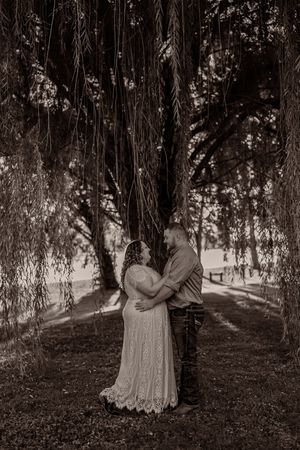 A romantic couple embraces under flowing willow tree branches in a dreamy black and white photograph.