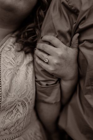 Close-up black and white photo showing hands with engagement ring against lace dress fabric.