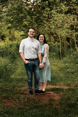 Casual engagement photo of couple standing together on forest path surrounded by green foliage.