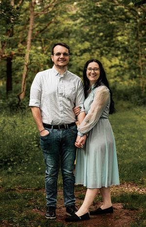 Couple in casual formal wear posing together on dirt path in forest setting.