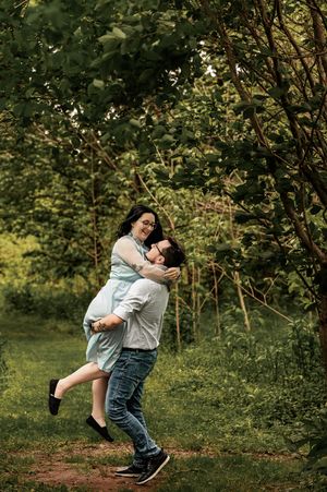 Playful engagement photo with man lifting woman in sage dress against lush green forest background.