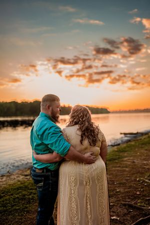 A couple embraces while watching a stunning orange sunset over a peaceful lake surrounded by trees.