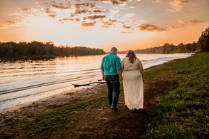 A couple walks along a riverbank at sunset with orange clouds reflecting on the water's surface.