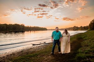 A couple stands together on a beach shore at sunset with orange clouds reflecting on the calm water.