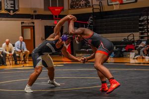 Two wrestlers compete in a match on a gymnasium mat while officials observe in the background.