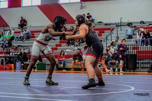 Two wrestlers compete in a high school wrestling match on a red mat while officials and spectators watch.