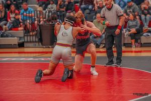 Two wrestlers compete on a red wrestling mat during an intense match while officials and spectators watch.