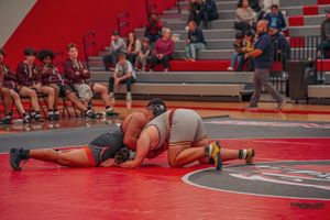 High school wrestling match on red mat as spectators watch from bleachers in gymnasium.