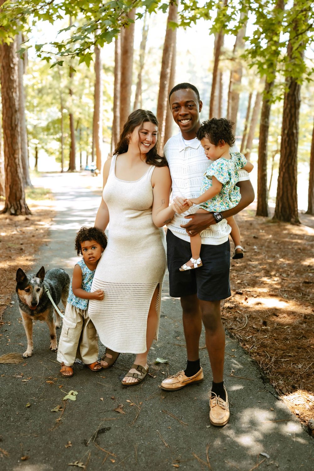 Family spending time together in a sunlit forest with dappled light filtering through tall pine trees on a dirt path.
