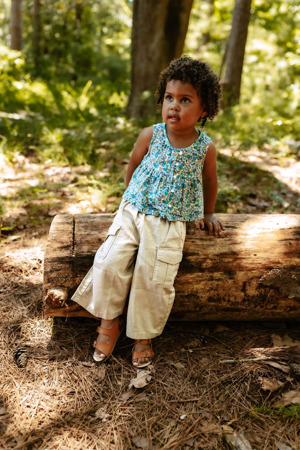 Young child in turquoise floral top and khaki cargo pants sits casually on fallen log in sunlit forest setting.