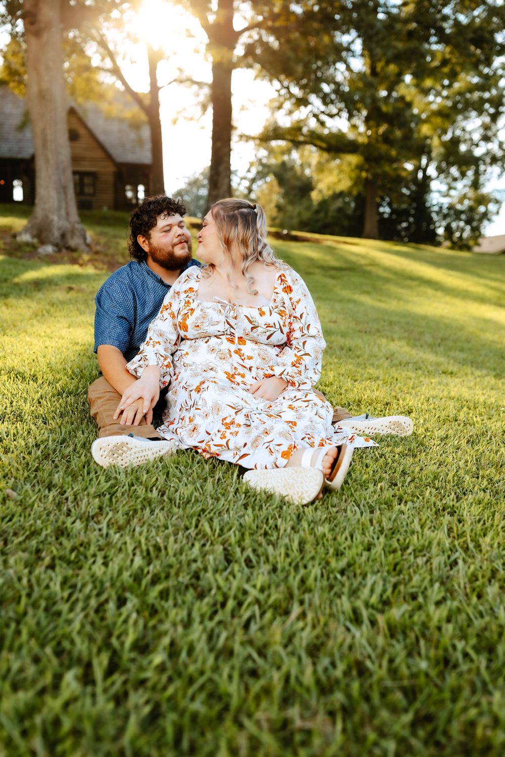A couple shares a romantic moment while lounging together on a sun-dappled grassy hillside during golden hour.