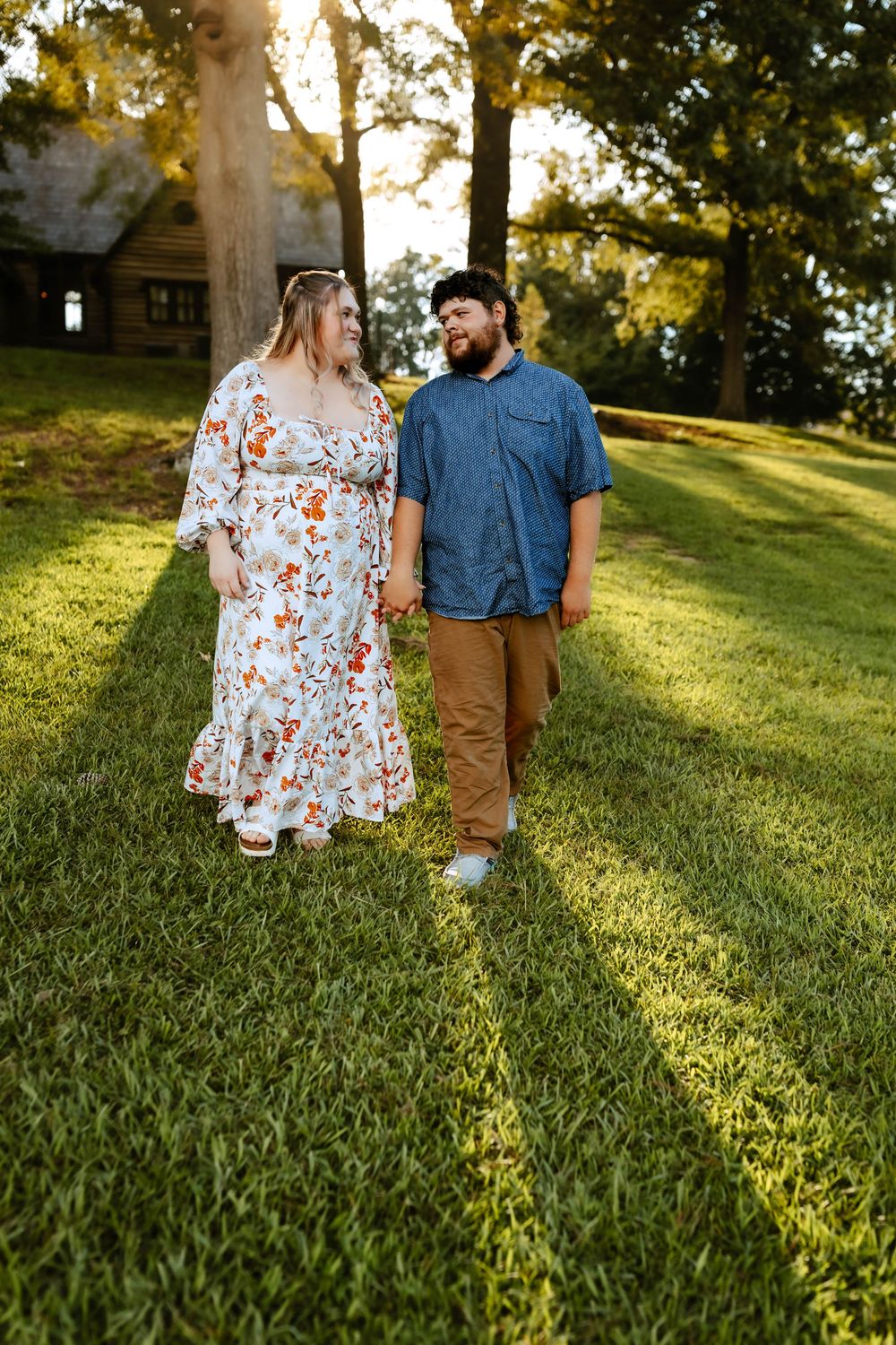 A couple walks together through a sunlit grassy hillside, dressed in a floral dress and denim shirt with khaki pants.