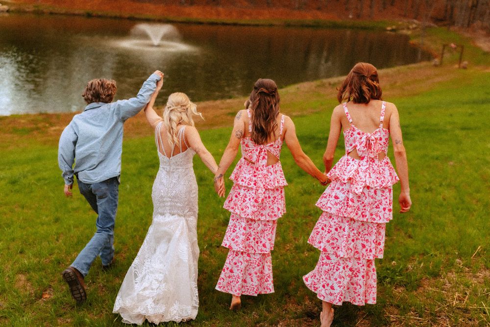 A group holding hands while walking along a lake shore in pink floral and white dresses on a summer day.