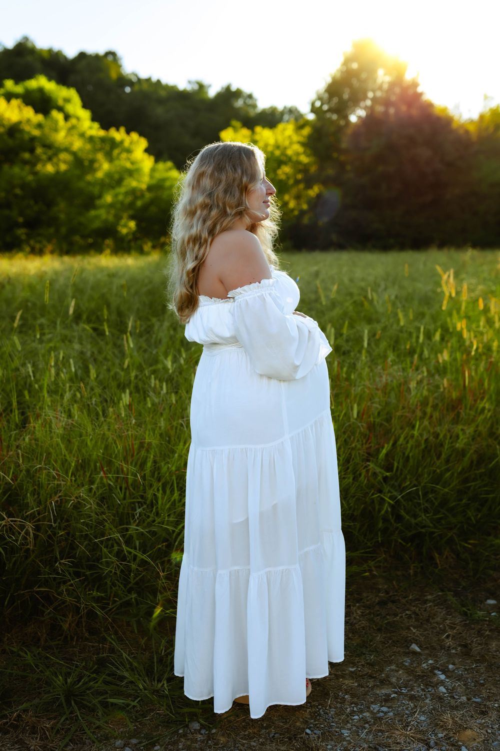 Two people in white dresses embrace in a sunlit meadow at golden hour as sunlight filters through trees.