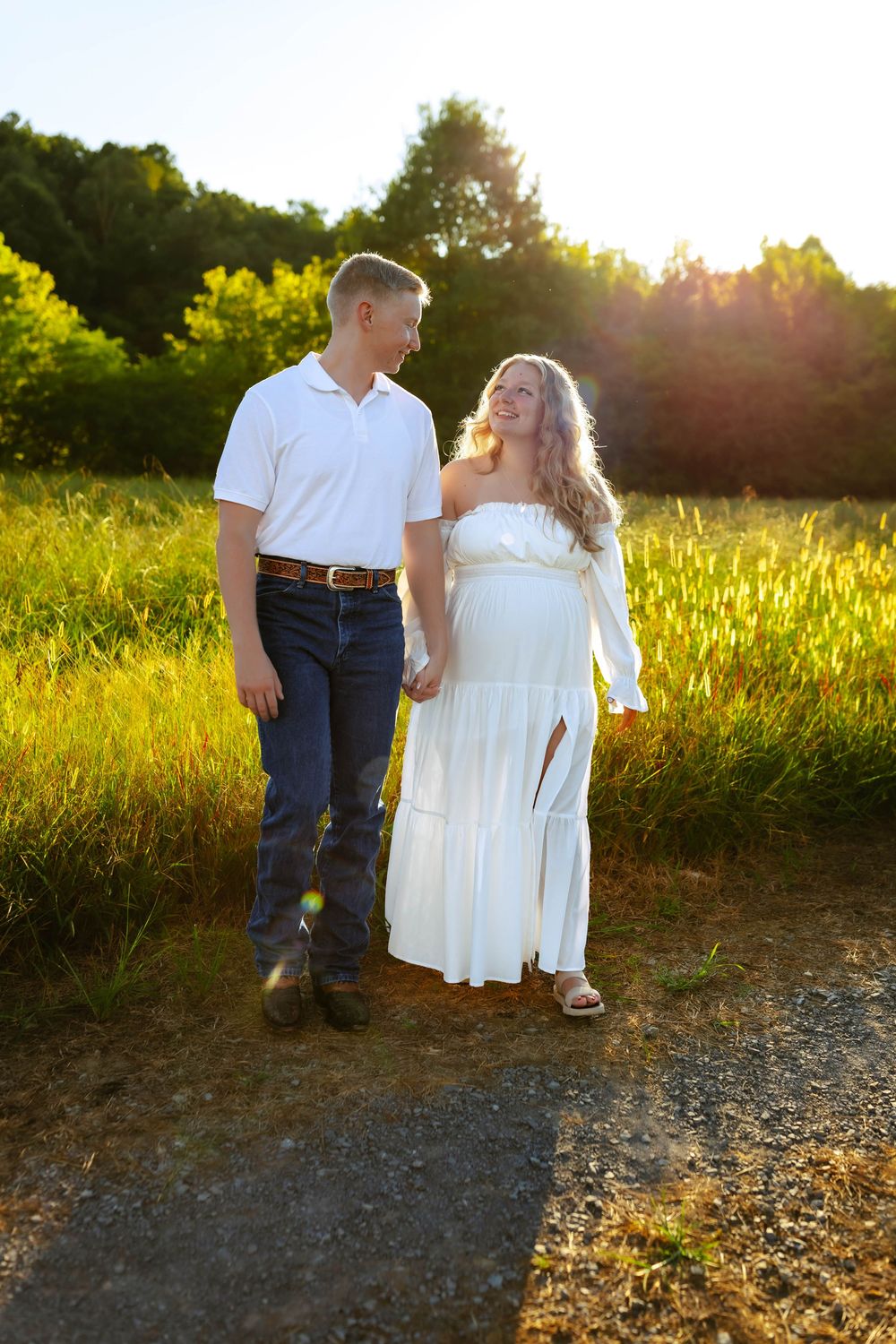 A couple holds hands while walking through a golden sunlit meadow at sunset wearing white and denim.