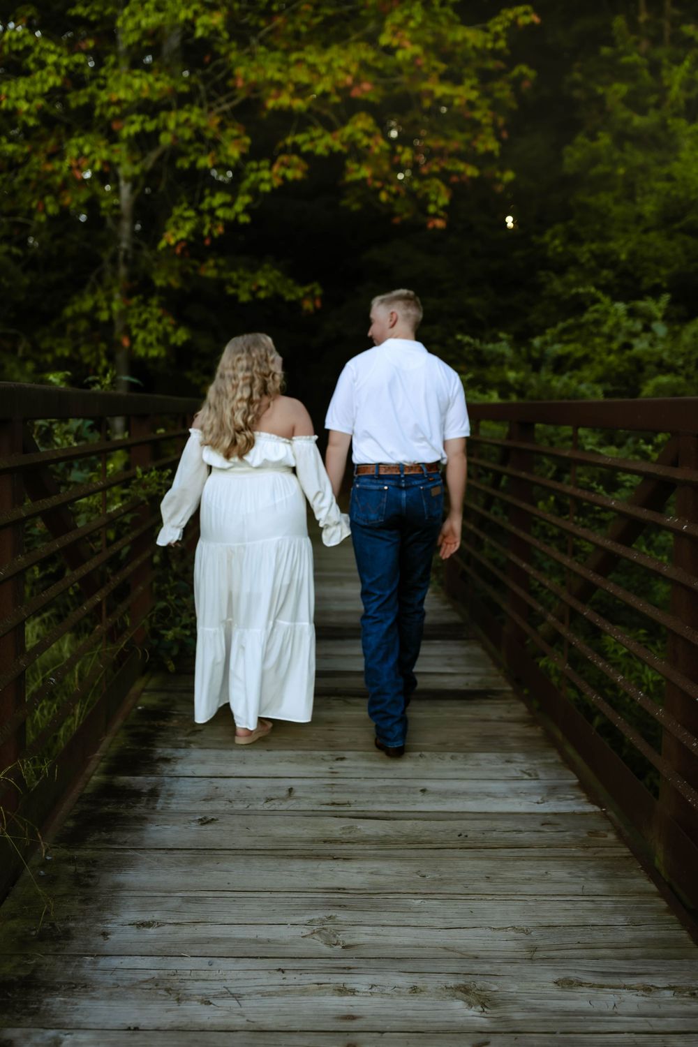 A couple in white dress and blue jeans walking away together on a rustic wooden bridge surrounded by green foliage.