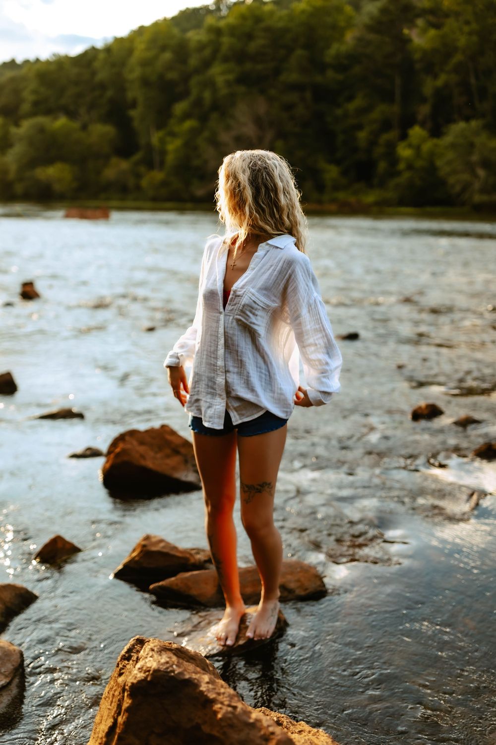 A person in a white shirt and shorts explores a rocky shoreline along a peaceful lake at sunset.