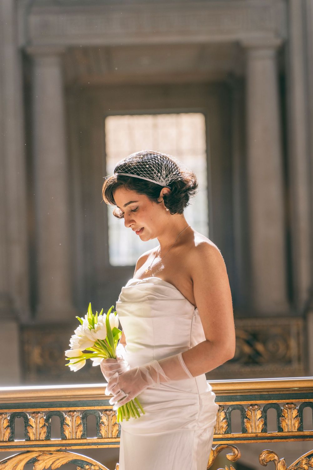 Elegant bride in strapless gown holds white tulip bouquet wearing a birdcage veil in a grand hall.