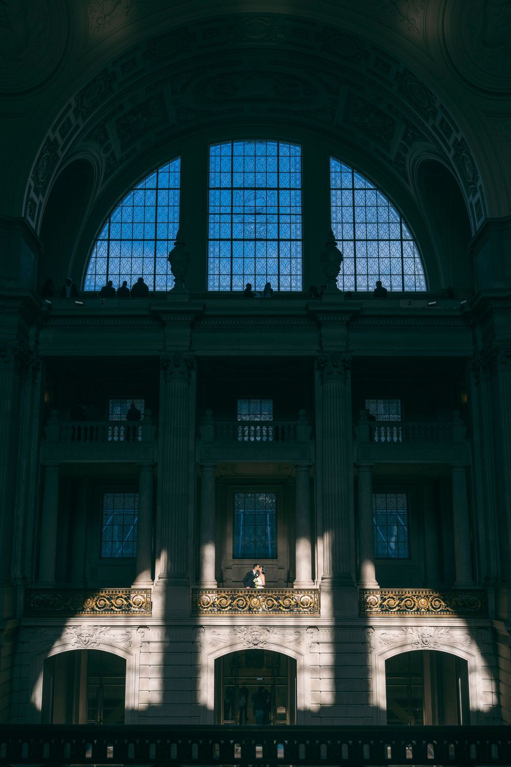 Lone bride stands on ornate gold balcony beneath a massive arched window in a grand civic building.
