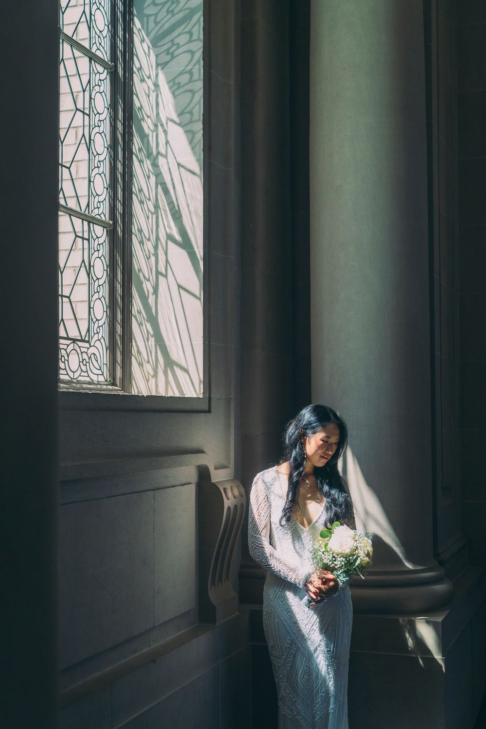 Bride in white lace gown holding bouquet by stained glass window in elegant sunlit interior.
