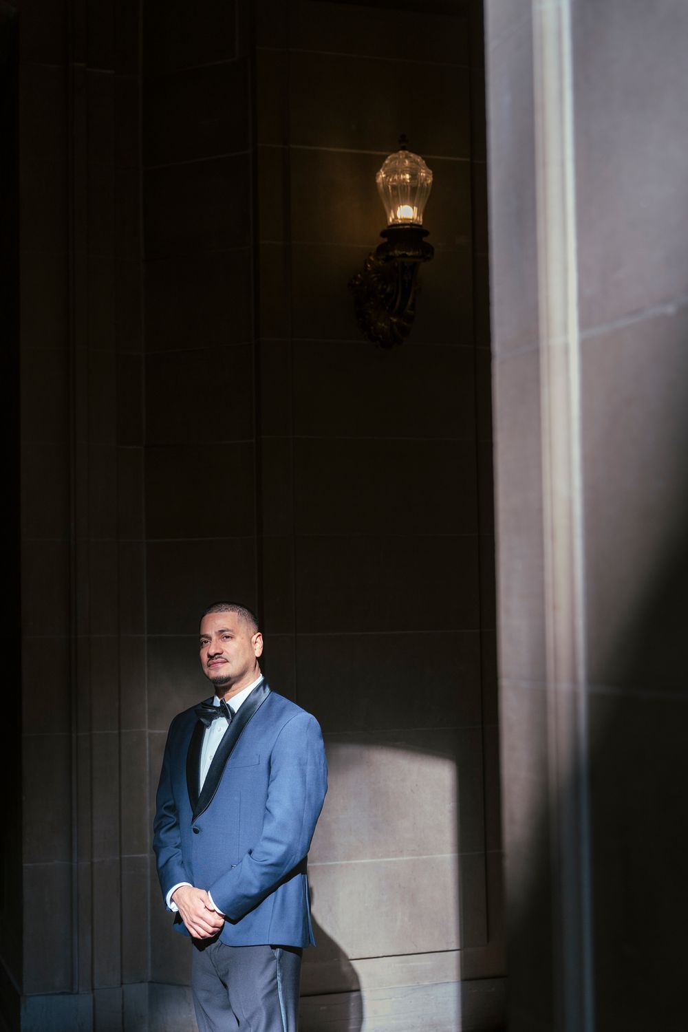 A dapper groom in a blue tuxedo and bow tie stands against a stone wall beneath a vintage wall sconce.