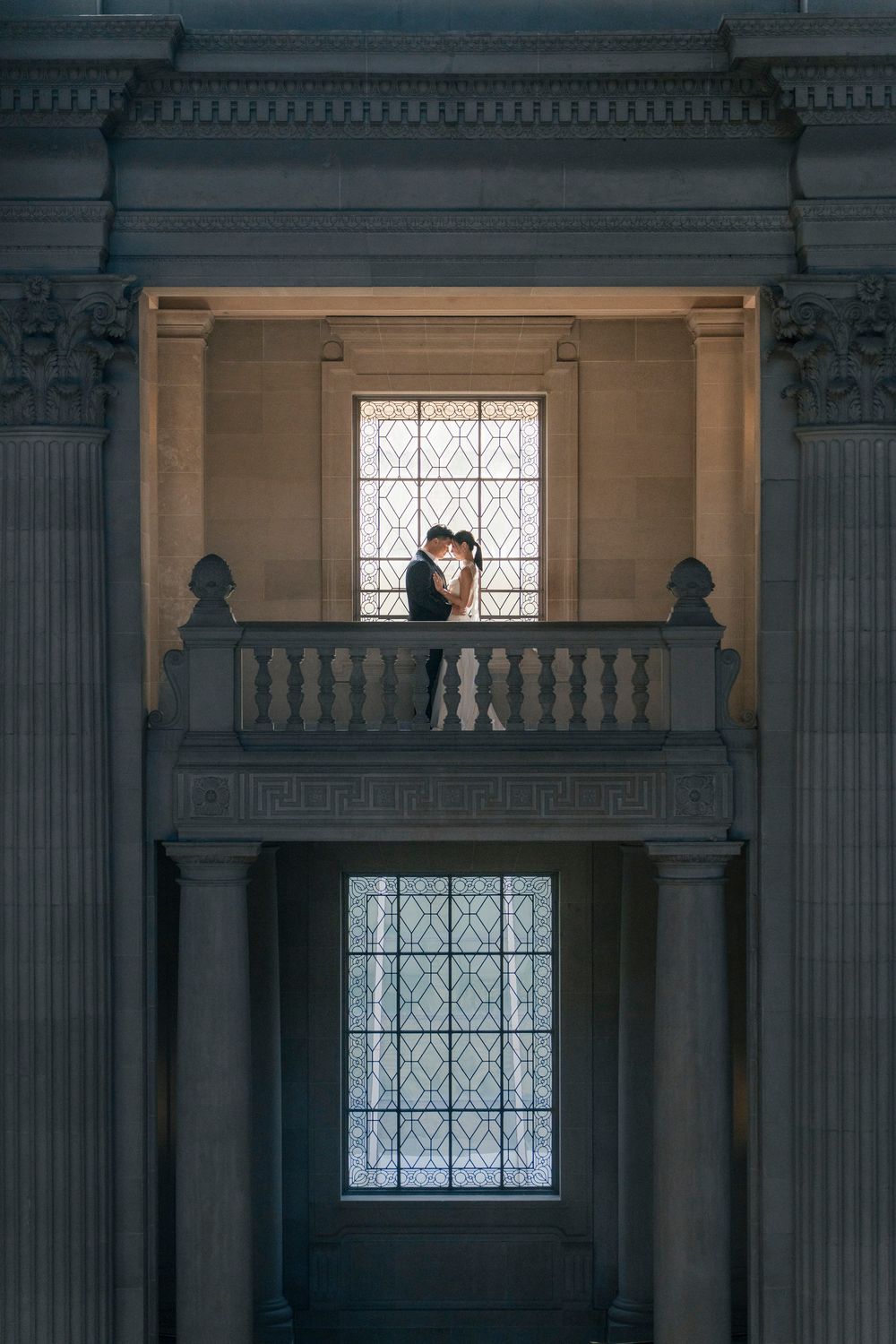 Couple embracing on ornate balcony with columns and leaded glass windows in elegant neoclassical building.