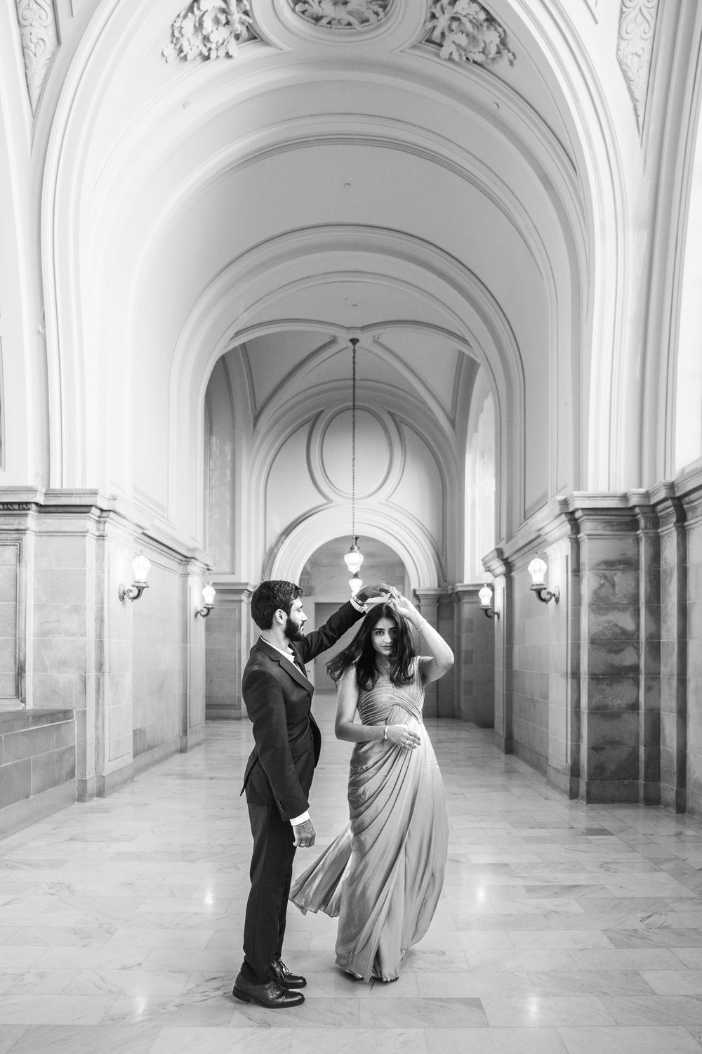 Black and white photo of a couple dancing elegantly in a grand arched hallway, man spinning woman in flowing gown.