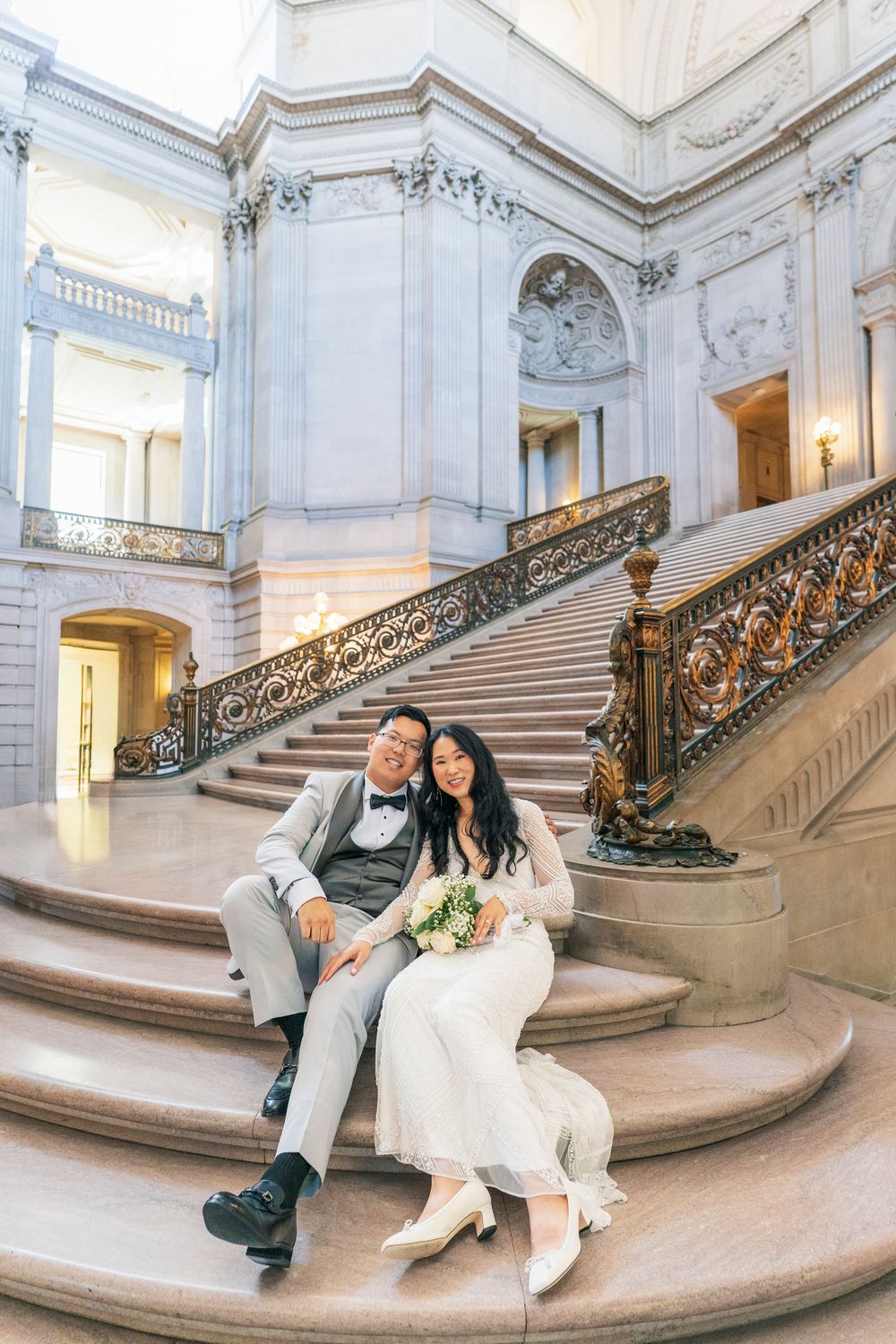 Bride and groom sitting together on grand marble staircase inside San Francisco City Hall on wedding day.