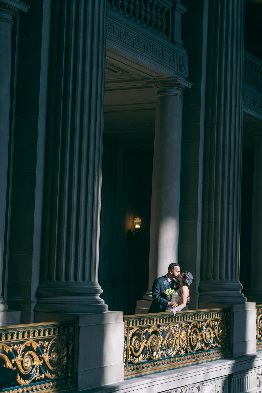 Couple sharing a romantic kiss on an ornate gold balcony between grand dark columns of a historic building.