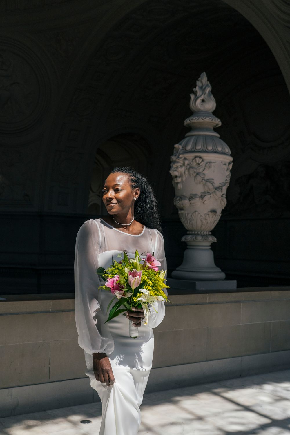 Elegant Black bride in white dress holding colorful bouquet, posing near ornate stone vase architecture.