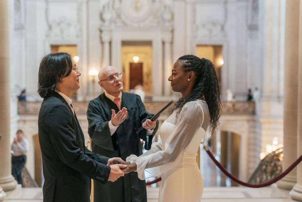 Couple exchanging vows at San Francisco City Hall wedding ceremony with officiant clapping in elegant rotunda.