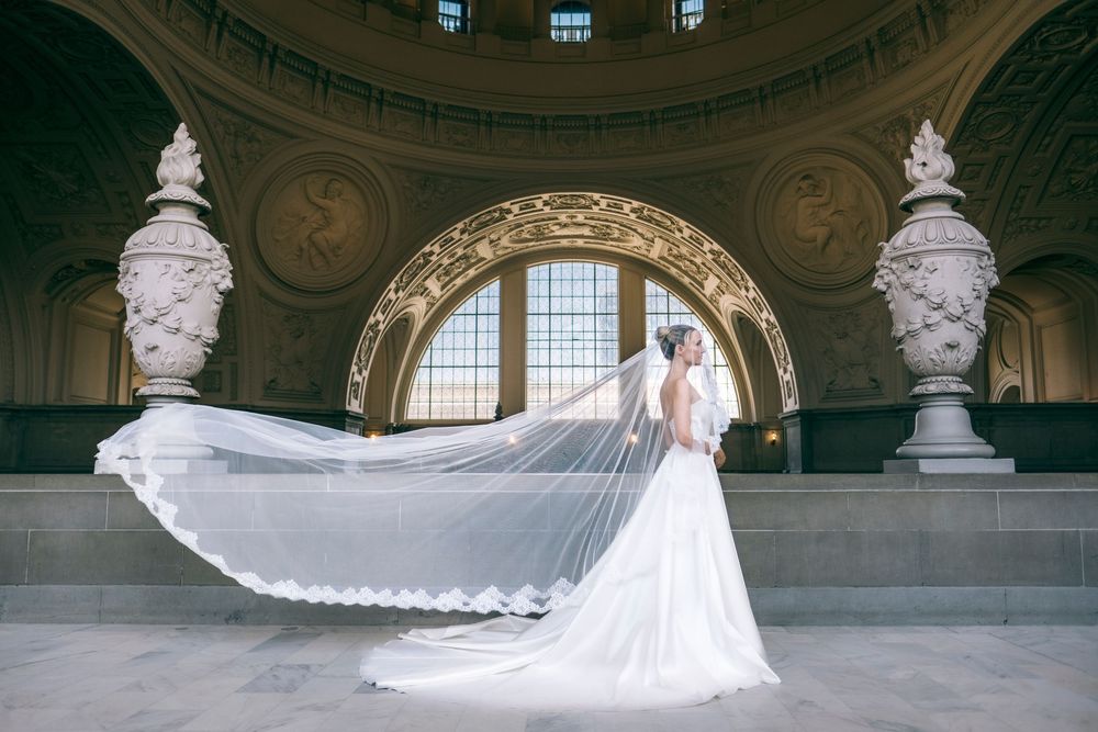 Bride in white gown with flowing cathedral veil in ornate domed hall with arched windows.