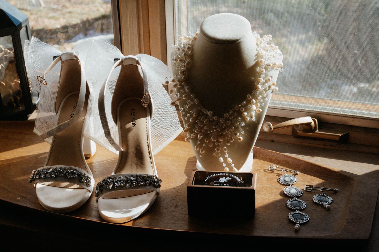White bridal shoes and elegant pearl necklace displayed on wooden table near window with jewelry accessories. Wedding details pictures.