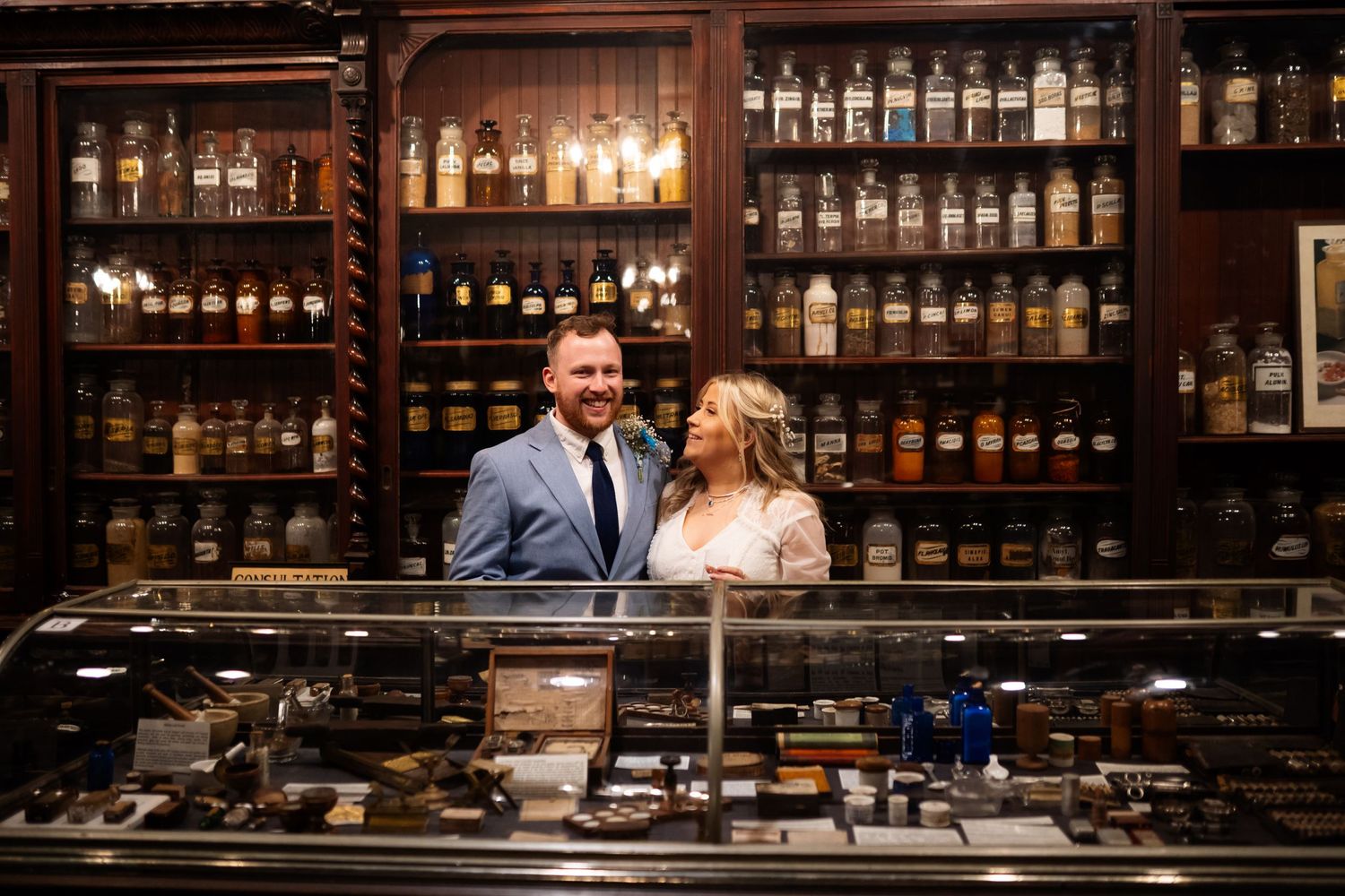 A couple stands near a bar counter with vintage apothecary bottles and jars displayed on wooden shelves behind them.