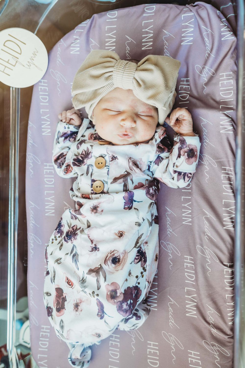Sleeping newborn wearing floral print sleeper and bow headband on purple patterned background.