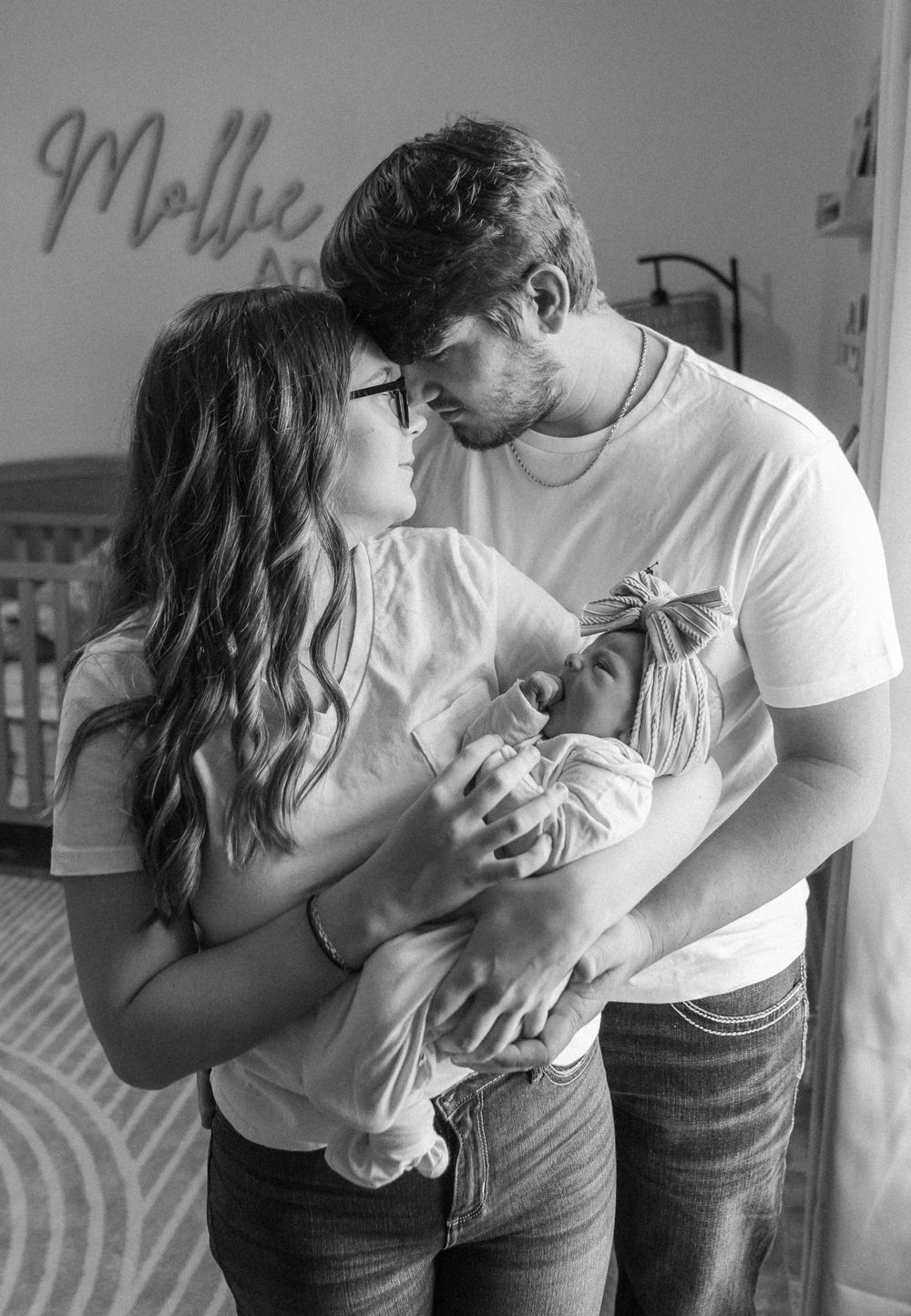 Black and white image of parents holding newborn baby in nursery room with name on wall.
