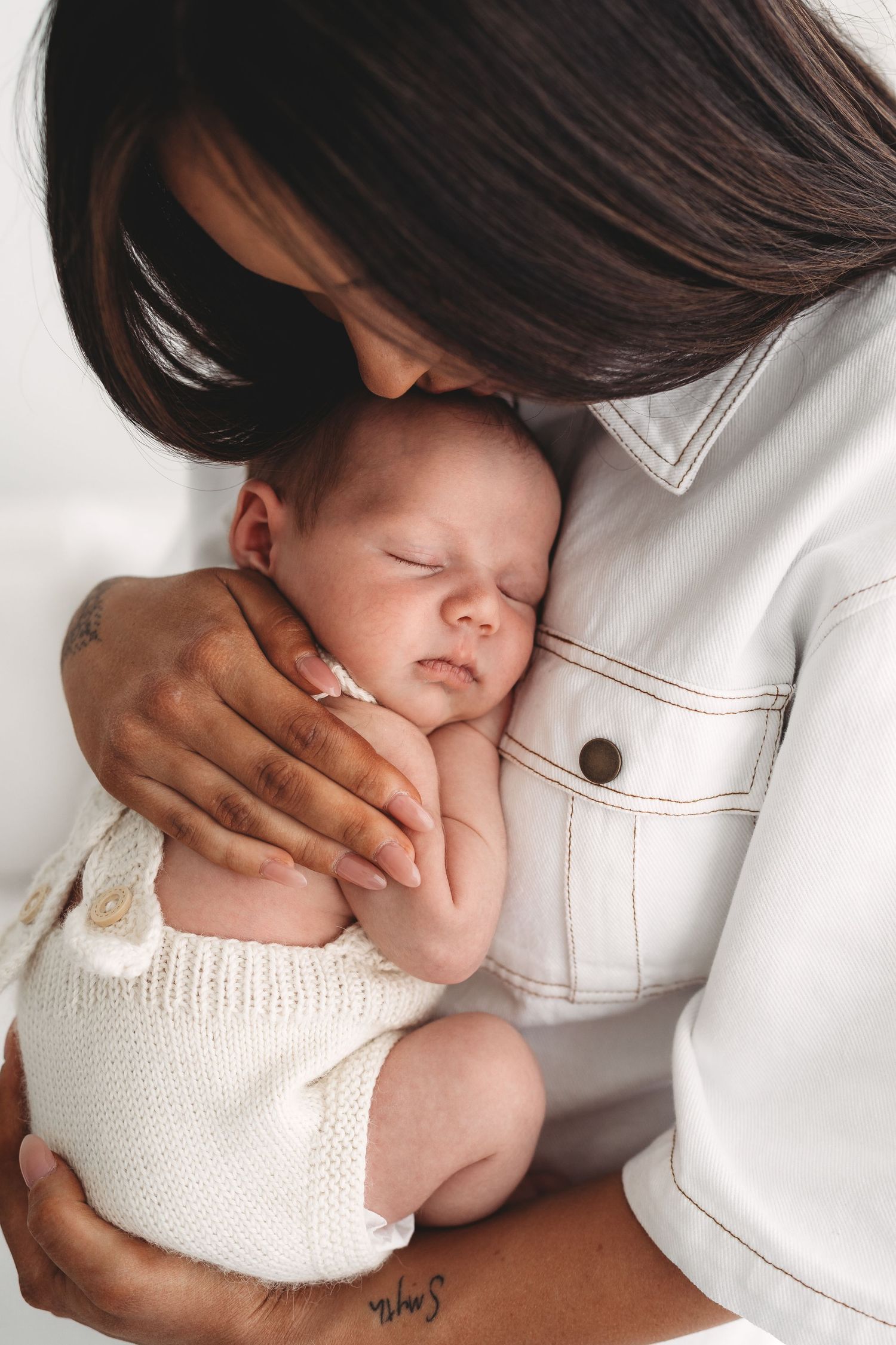 Mother with dark hair gently kissing her sleeping newborn baby dressed in cream knit.