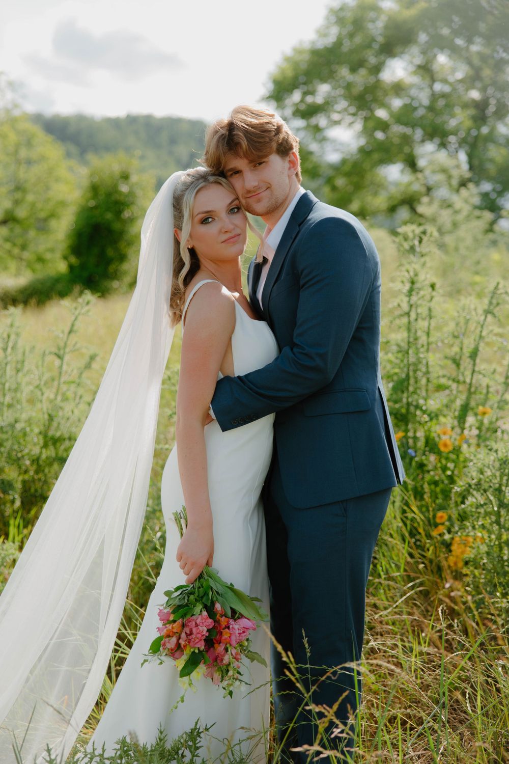 A romantic wedding portrait of a couple embracing in a sunlit meadow with wildflowers and a flowing white veil.