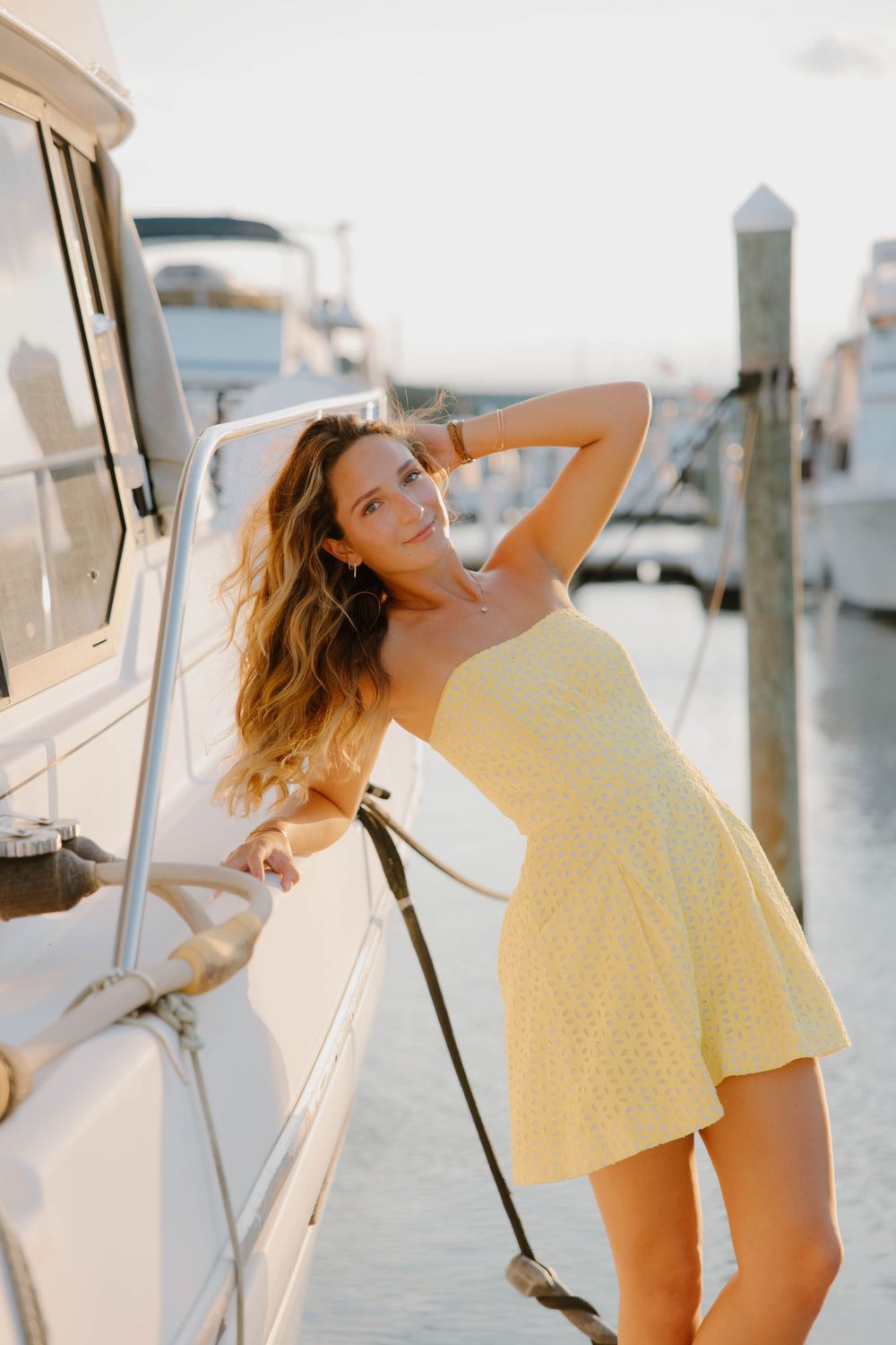 Woman in yellow sundress poses elegantly against boat at marina during sunset.