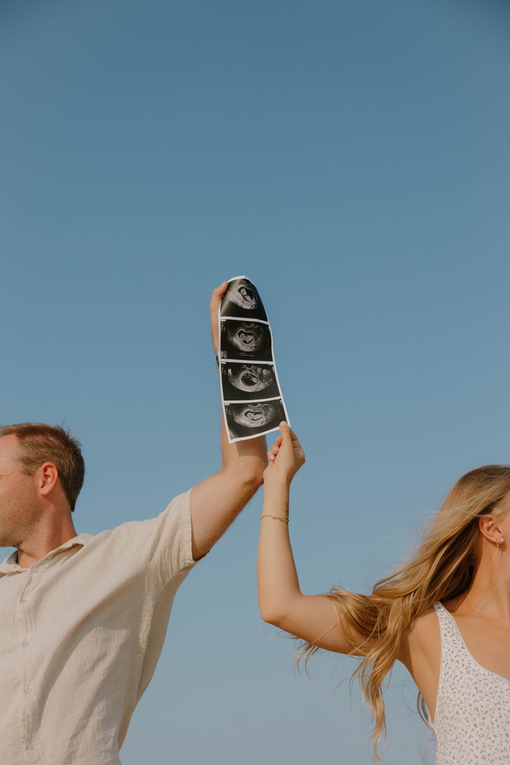 A couple holds up ultrasound photos against a bright blue sky.